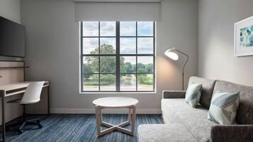 Una habitación con escritorio, una silla, sofá, mesa, lámpara y ventana con vista a la vegetación.