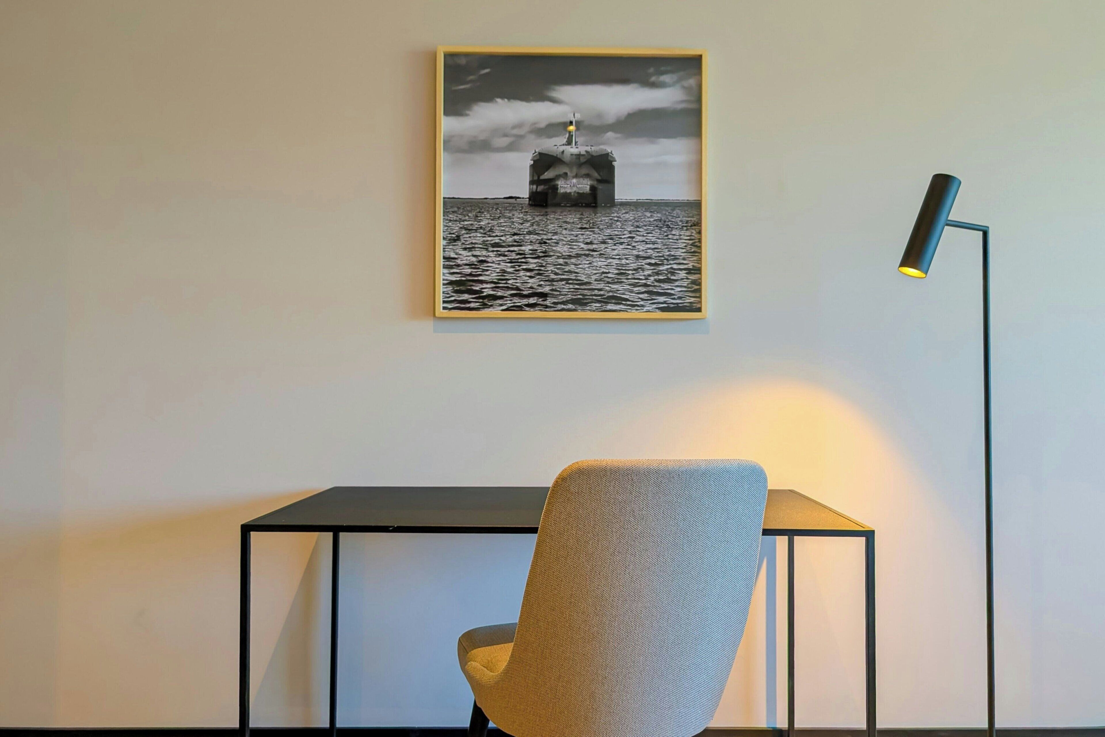 A framed photograph of a ship on the water, a desk with a chair, and a floor lamp.