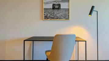 Uma fotografia emoldurada de um navio na água, uma escrivaninha com uma cadeira e um abajur.