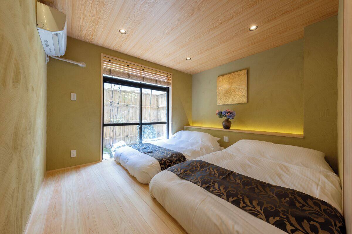 A bedroom with two beds, a window with blinds, a wall-mounted air conditioner, and a decorative vase on a shelf.