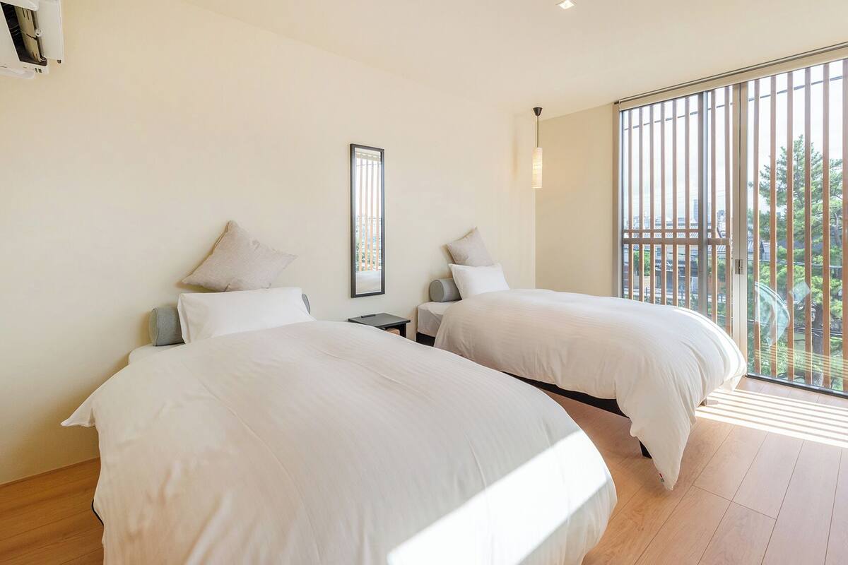 A bedroom with two beds, a window with blinds, a bedside table, and a wall-mounted air conditioning unit.