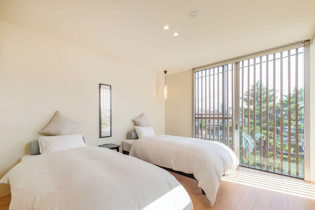 A modern hotel room with two beds, a large window with vertical blinds, and a view of the cityscape.