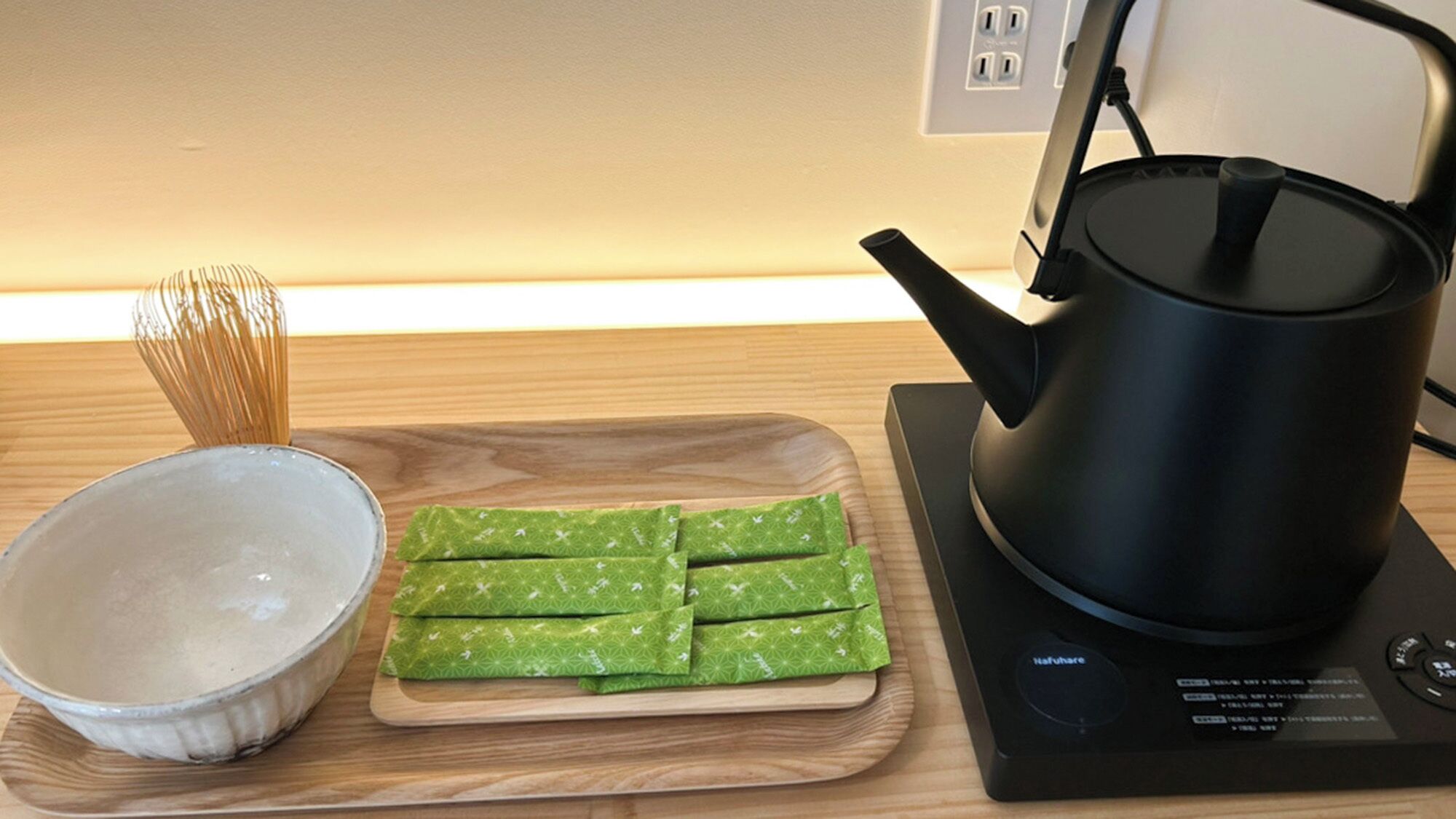 Un plan de travail de cuisine avec une bouilloire électrique noire, une planche à découper en bois ornée de feuilles vertes et un saladier blanc.