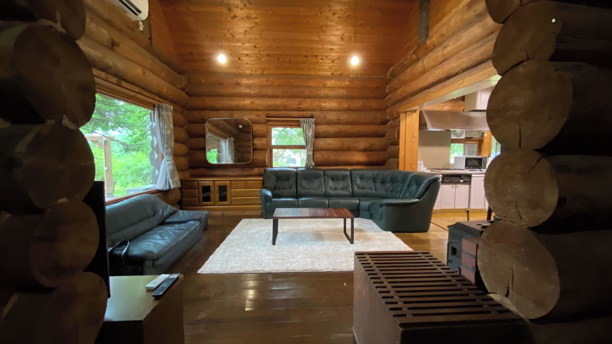 A log cabin interior with a sofa, a coffee table, a kitchen area, and a large window.