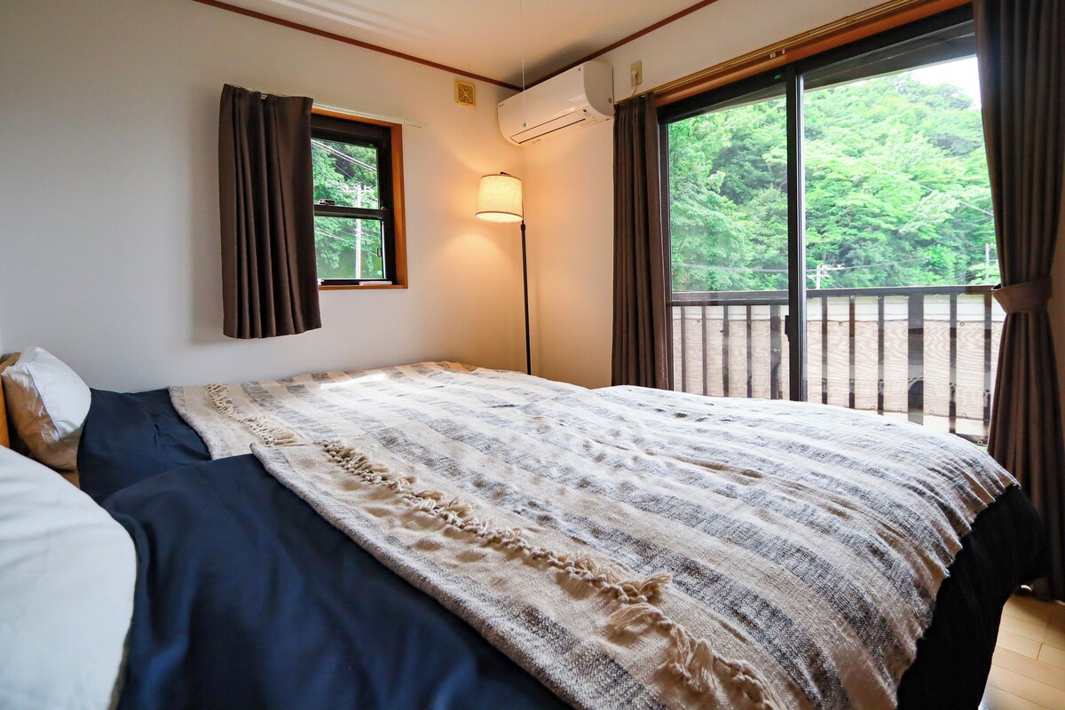 A bedroom with a bed, a window, a lamp, and a view of greenery.