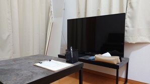 Ein Zimmer mit einem Flachbildfernseher auf einem Standfuß, einem kleinen Tisch mit einer Aktenablage, einer Taschentuchbox und einem schwarzen Netzkörbchen.