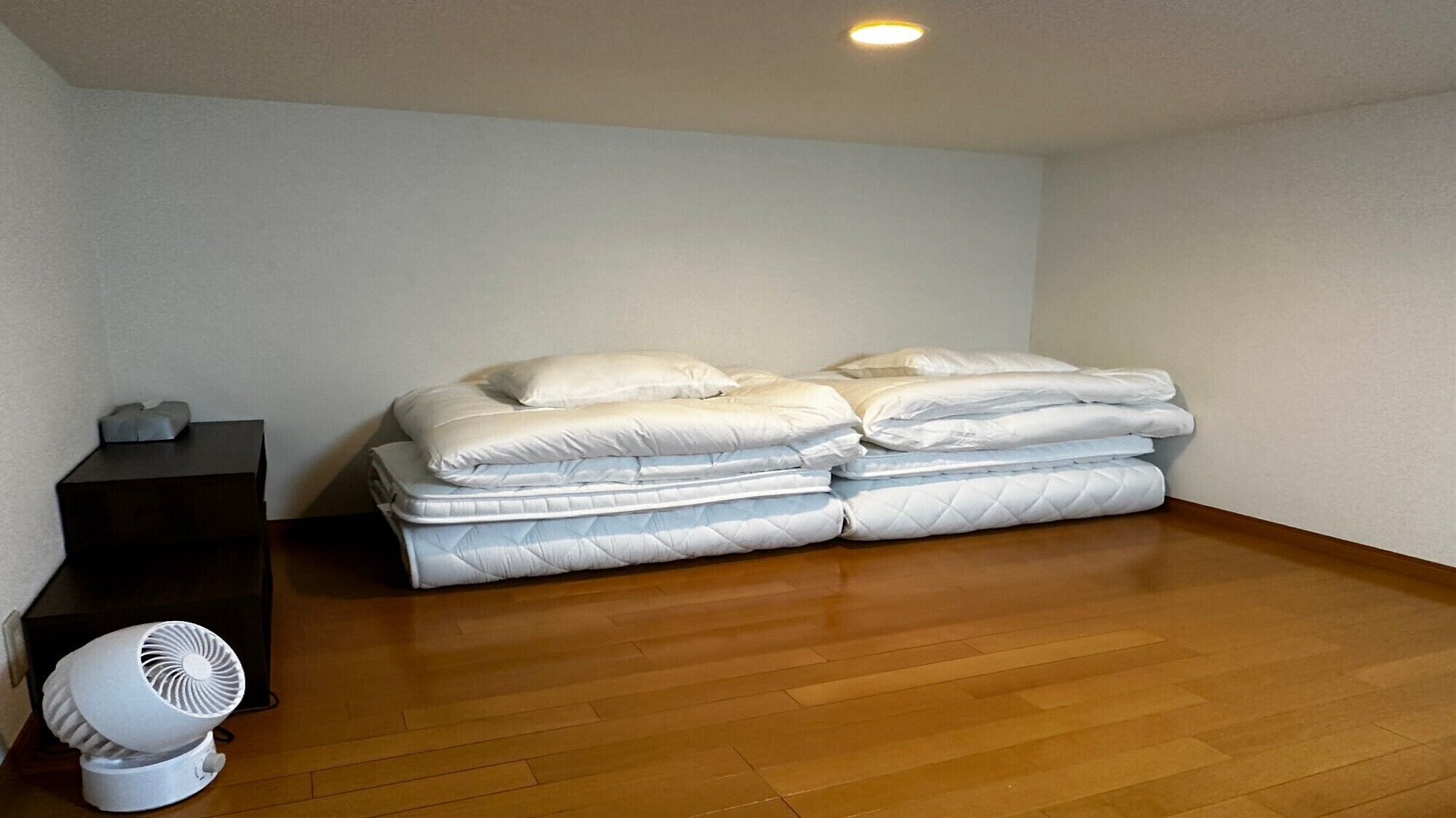Una habitación con piso de madera, una cama con ropa de cama blanca, una mesita con un ventilador y una pared blanca.
