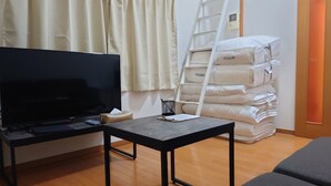 Ein Zimmer mit Fernseher, einem kleinen Tisch, einer Hockerleiter und einem Stapel Matratzen.