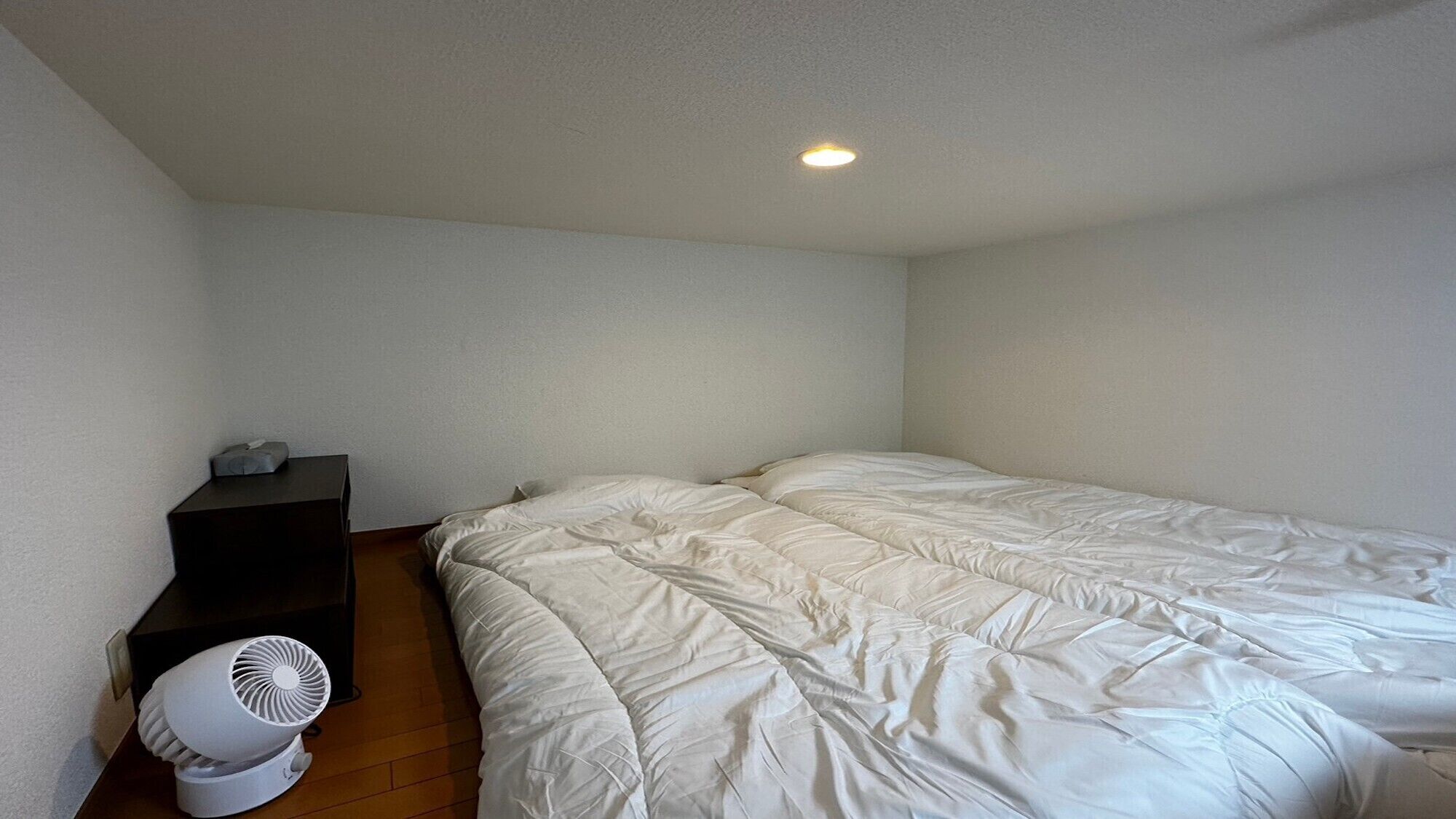 A bedroom with a bed, a small shelf, and a white fan.