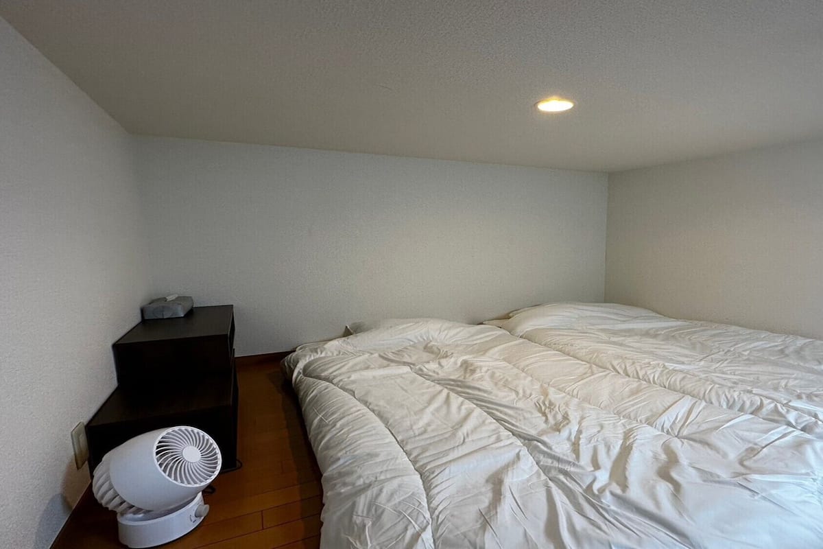 A bedroom with a bed, a small shelf, and a white fan.