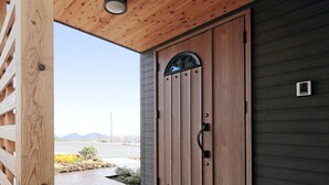 A wooden door with a handle and a window above it, set in a building with a sloped roof and a small outdoor area with a paved path and a few plants.