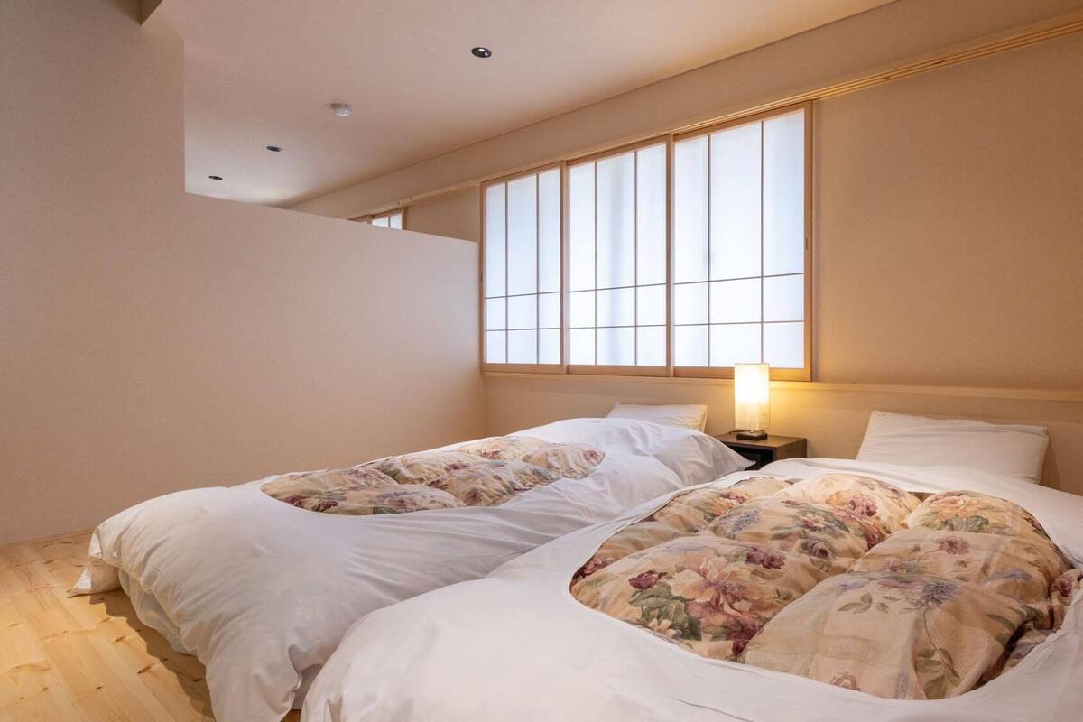 Ein Schlafzimmer mit zwei Betten, einem Fenster mit Jalousien, einer Nachttischlampe und Holzboden.