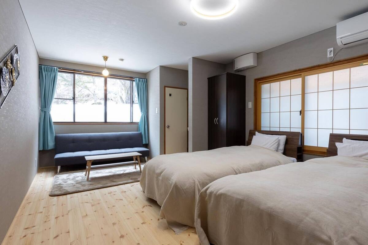 Ein Hotelzimmer mit zwei Betten, einer Bank, einem Fenster mit Vorhängen und einer Klimaanlage.