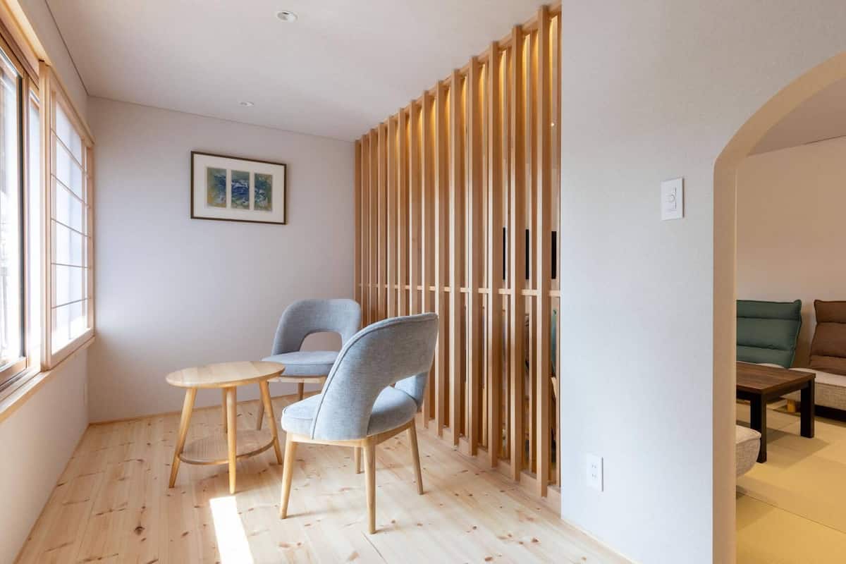 Ein Zimmer mit Holzboden, einem Sessel, einem kleinen runden Tisch, einer Couch und einem Raumteiler mit vertikalen Lamellen.