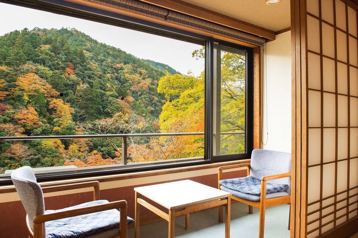 A room with a view of a forested hillside, a tatami mat wall, and a traditional Japanese-style table and chairs.