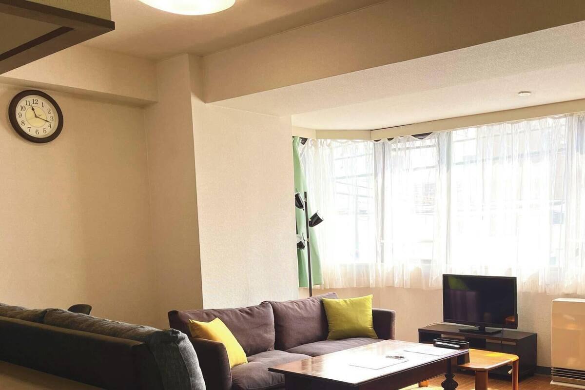 Uma sala de estar com sofá, mesa de centro, televisão e uma pequena mesa. Há um relógio na parede e uma janela com cortinas.