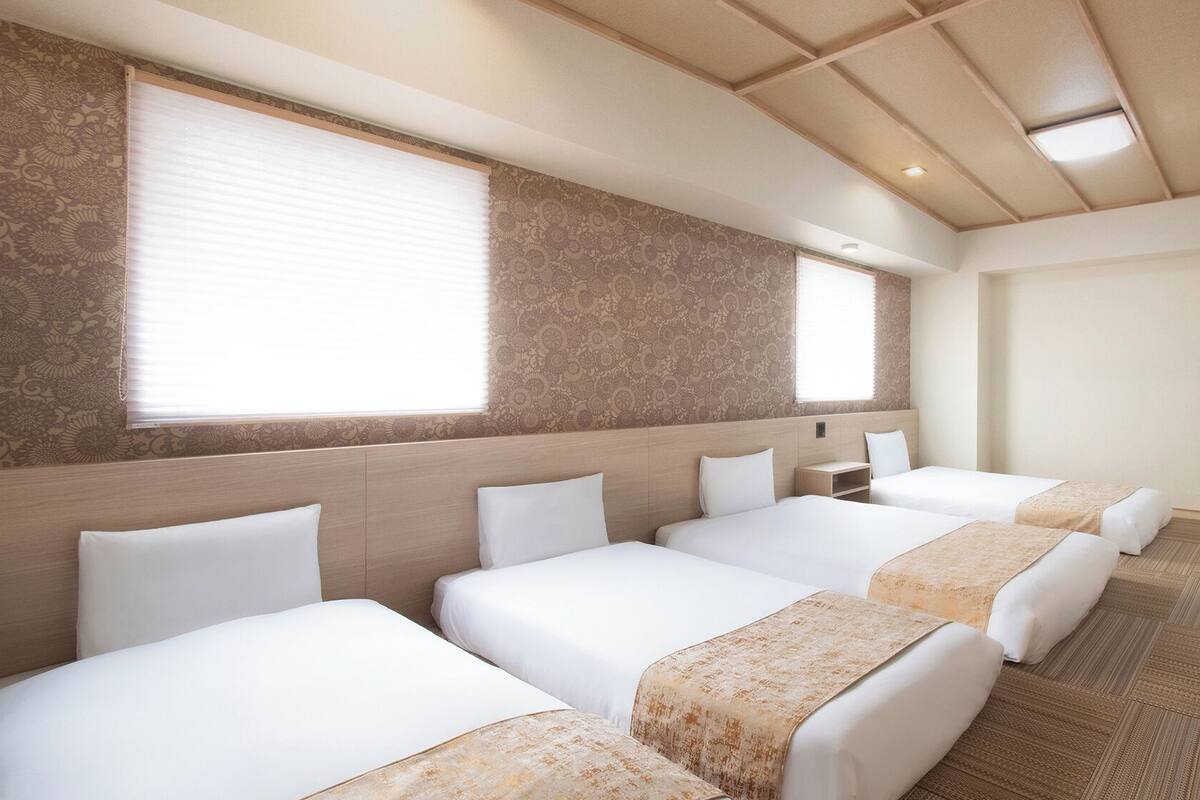 A hotel room with four single beds, a large window with blinds, a patterned wall, and a flat-screen TV mounted on the wall.