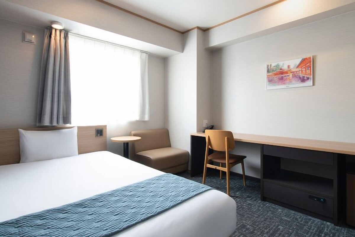 Ett hotellrum med en säng, ett skrivbord, en stol, ett litet bord och en tavla på väggen.