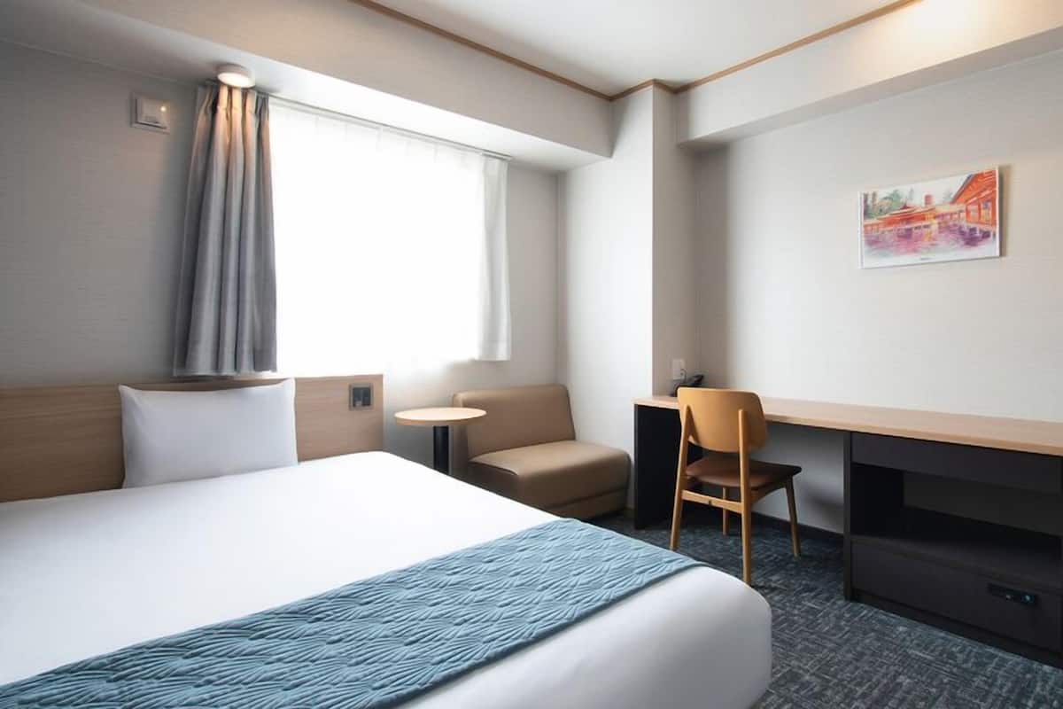 A hotel room with a bed, a desk, a chair, a small table, and a painting on the wall.
