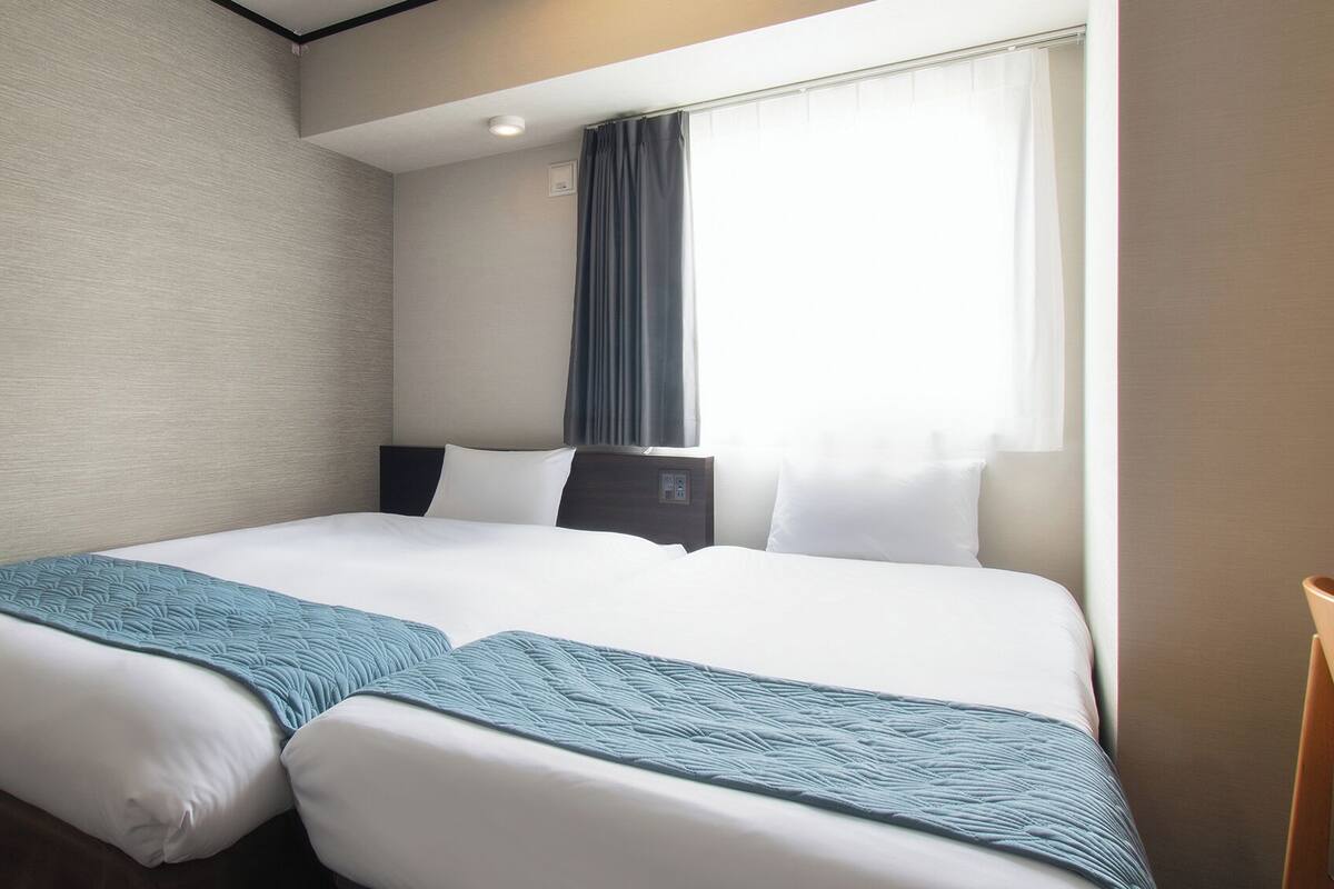 A hotel room with two beds, a window with curtains, a bedside table, and a chair.
