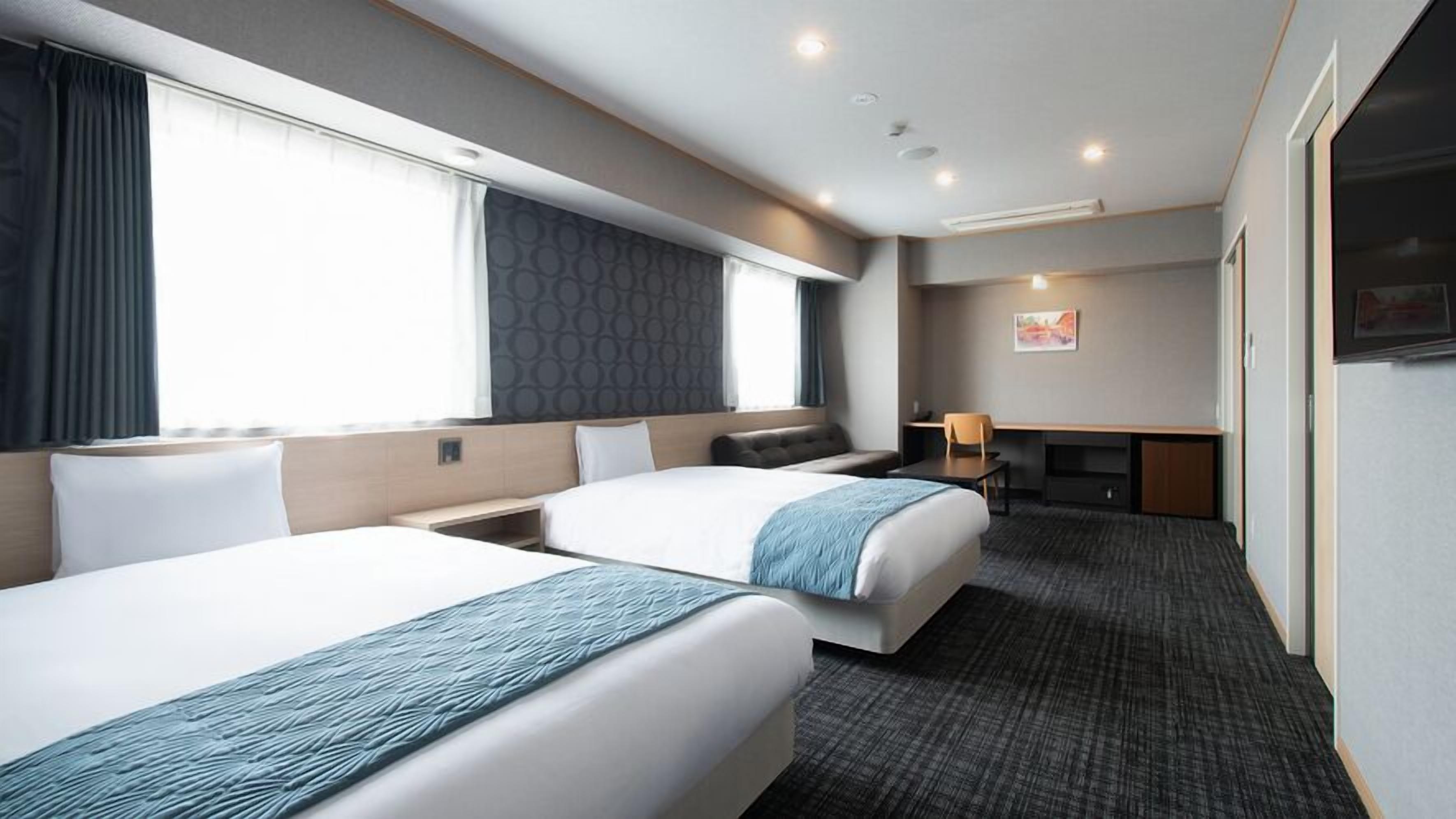 Ein Hotelzimmer mit zwei Betten, einem Schreibtisch, einem Sessel, einem Fernseher und einem großen Fenster.