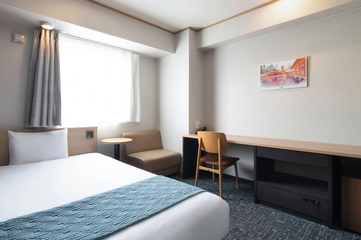 A hotel room with a bed, a desk, a chair, a small table, and a painting on the wall.