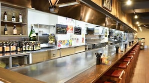 A bar area with shelves of bottles, a stainless steel counter, and a wooden bar stool seating area. - Vessel Hotel Hiroshima Heiwa Odori (Hiroshima)
