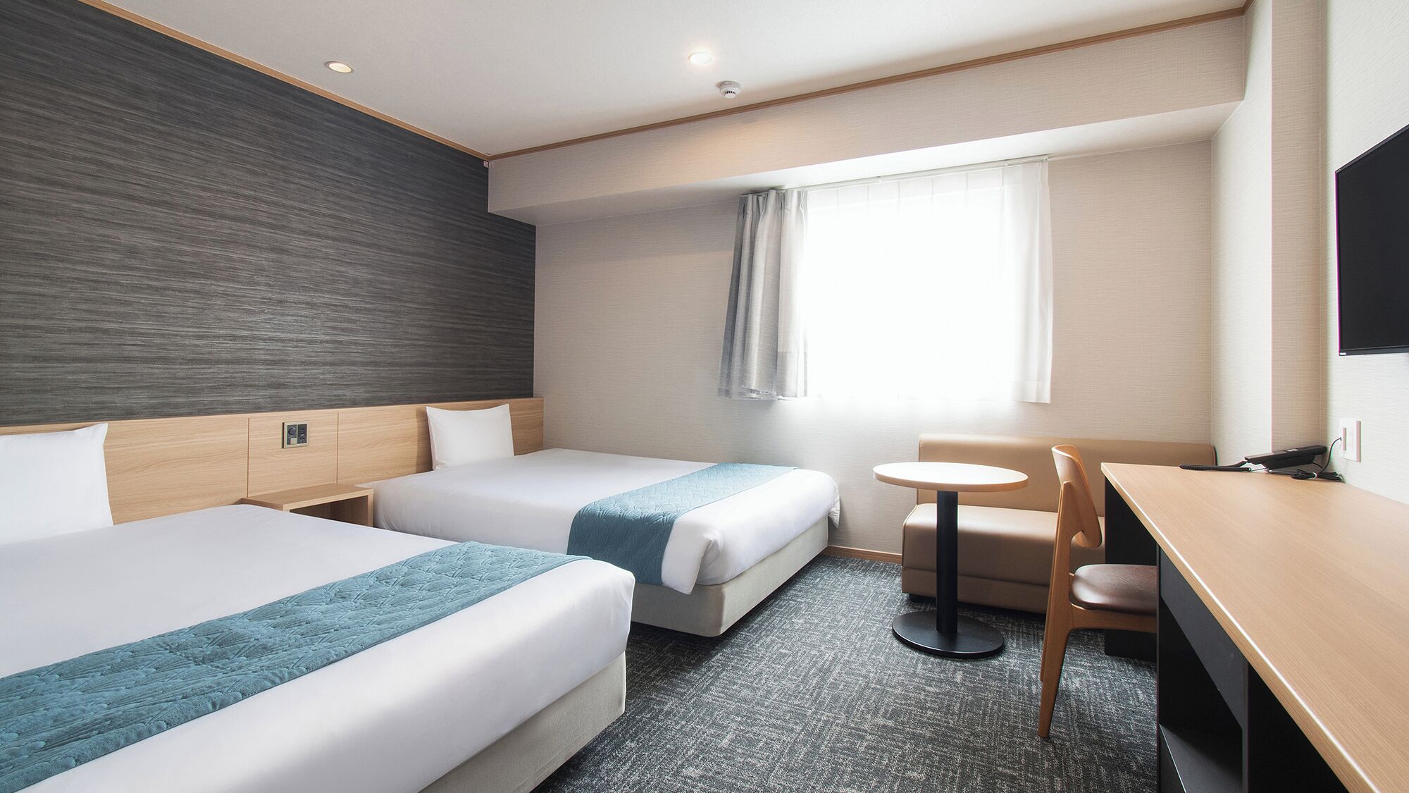Ein Hotelzimmer mit zwei Betten, einem Schreibtisch, einem Stuhl, einem kleinen Tisch und einem Fenster mit Vorhängen.