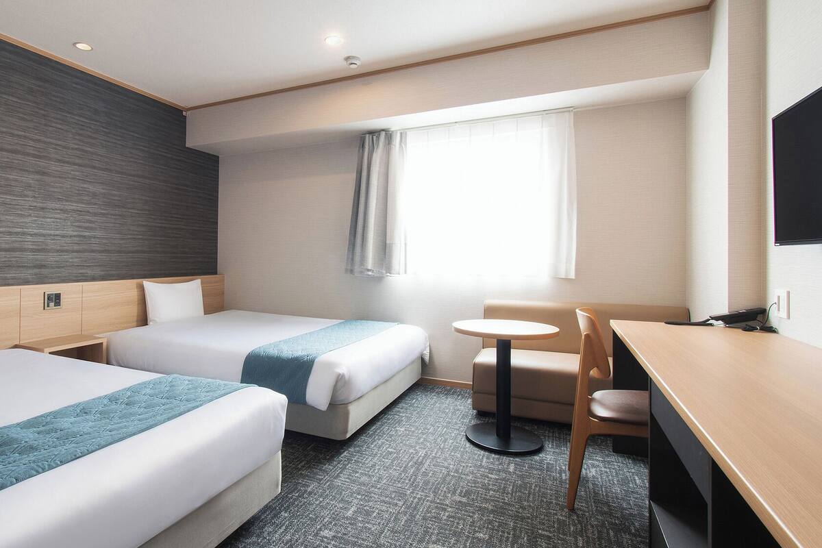A hotel room with two beds, a desk, a chair, a small table, and a window with curtains.