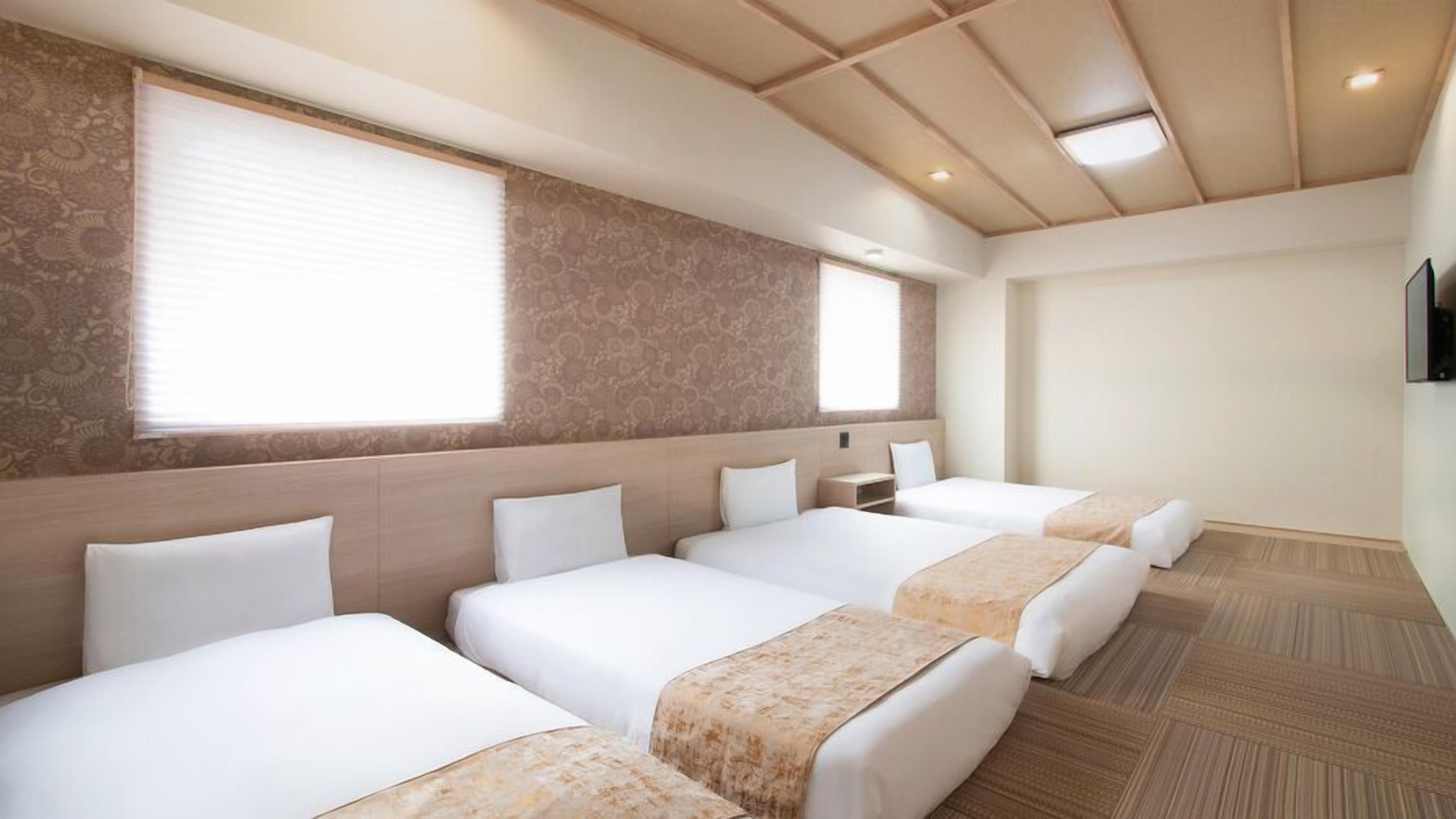 Ein Hotelzimmer mit vier Betten, einem großen Fenster, Wandtapete mit Muster und Holzboden.