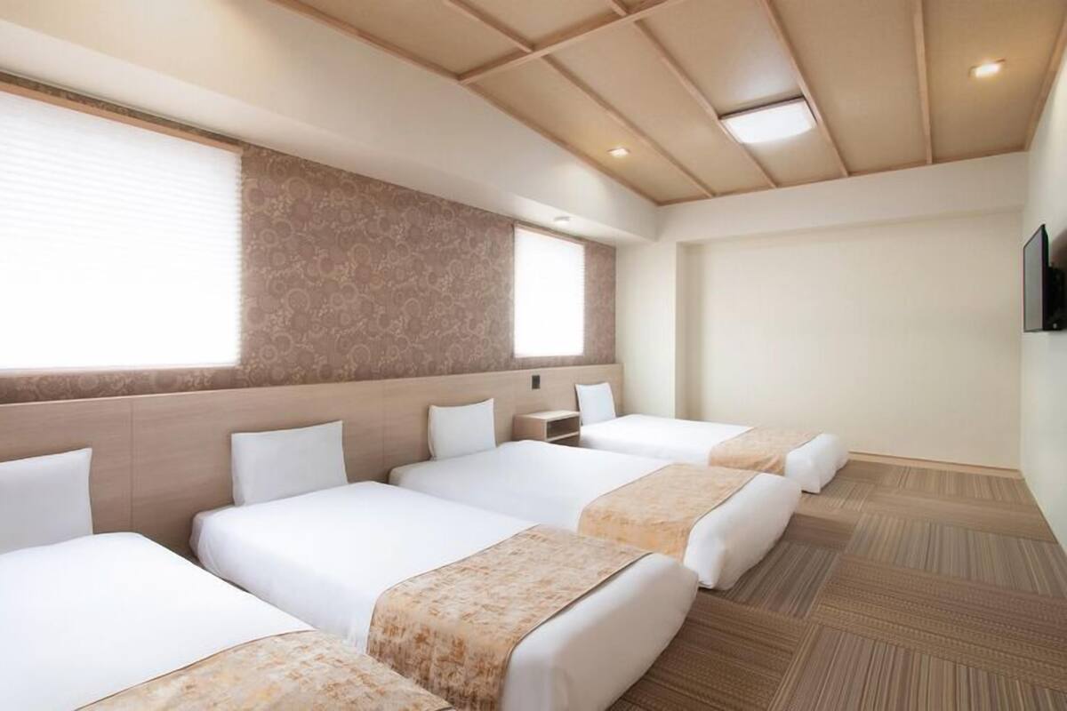A hotel room with four beds, a large window, patterned wallpaper, and a wooden floor.