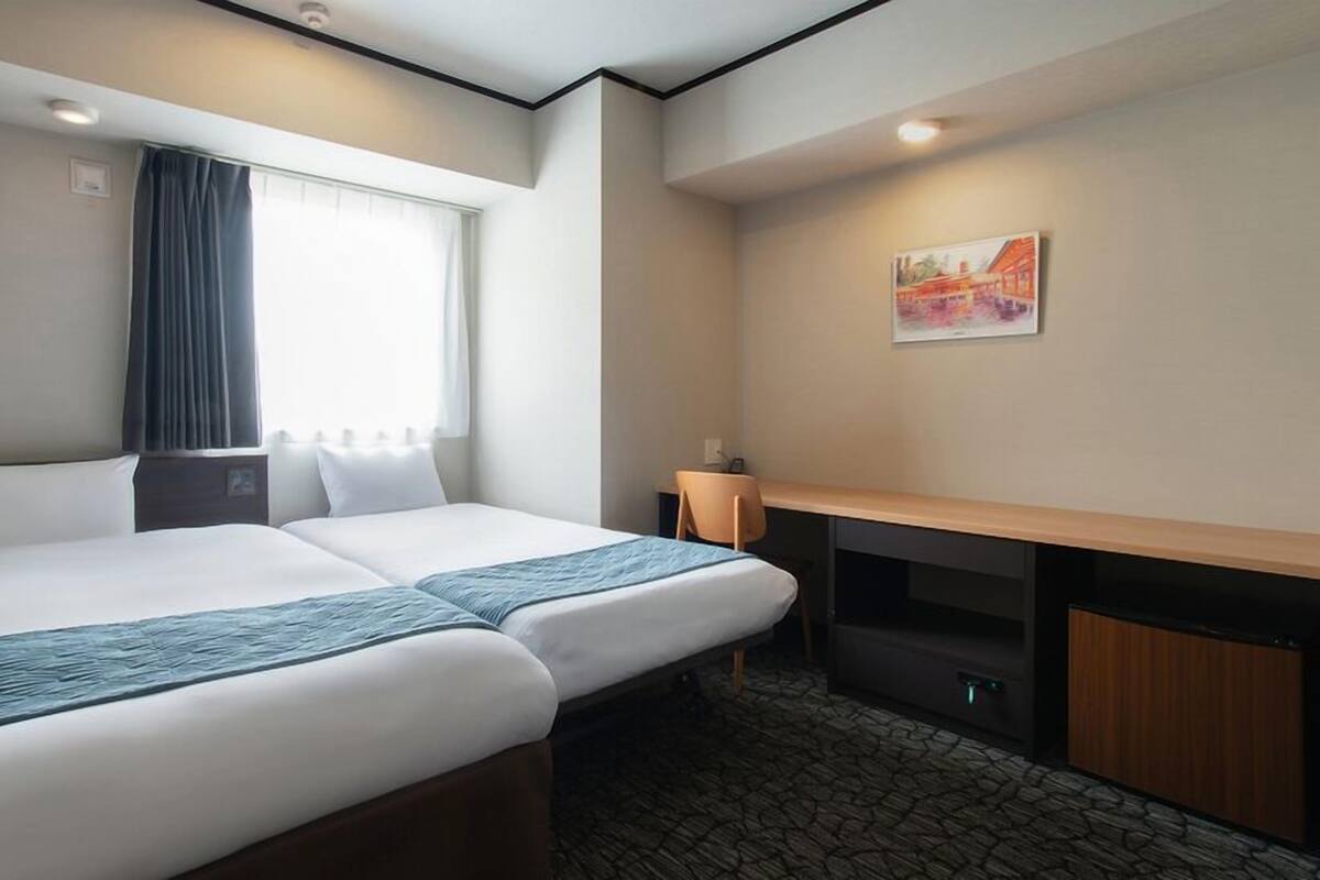 A hotel room with two beds, a desk, a chair, a window with curtains, a painting on the wall, and a stone-patterned floor.