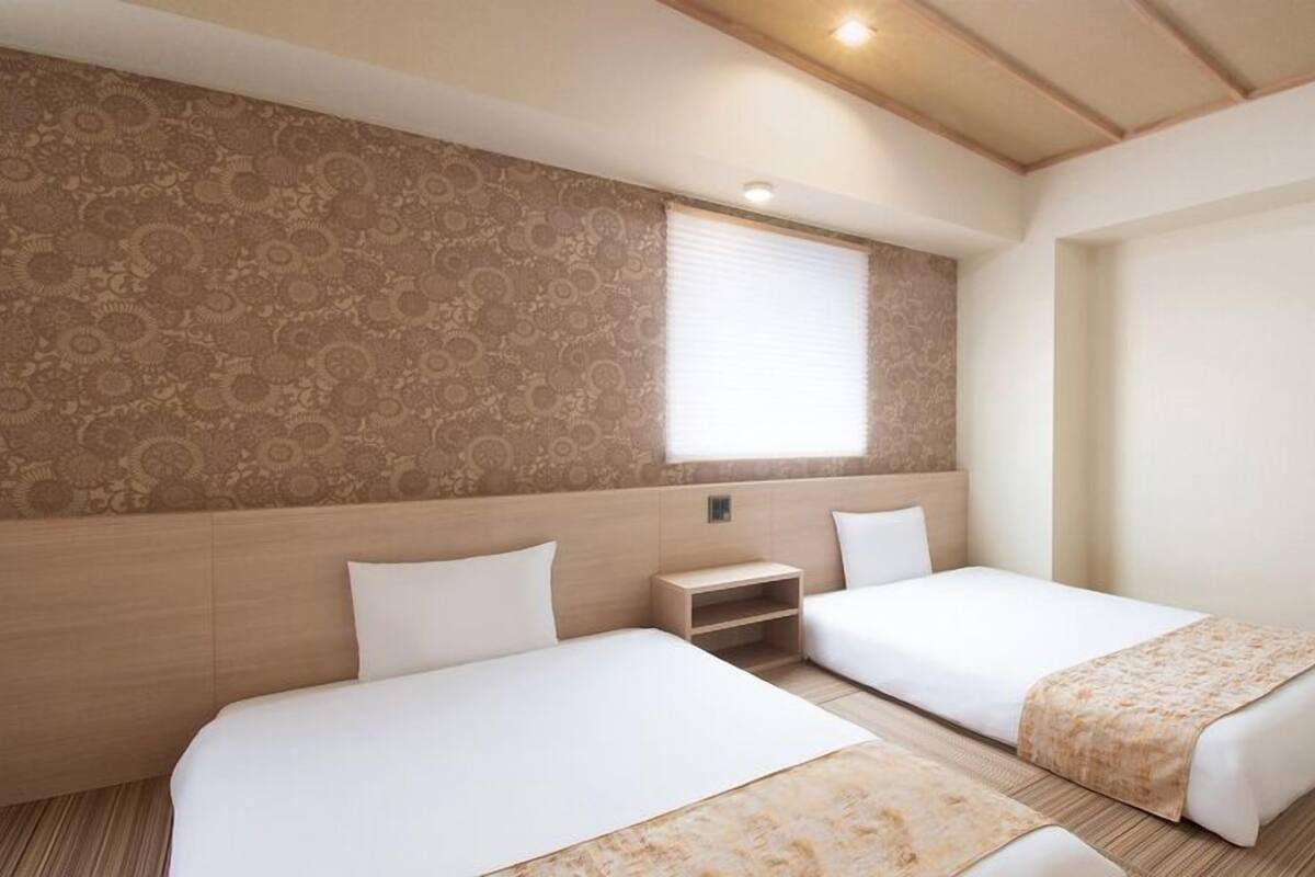 A hotel room with two beds, a small bedside table, a window with blinds, and a patterned wall.