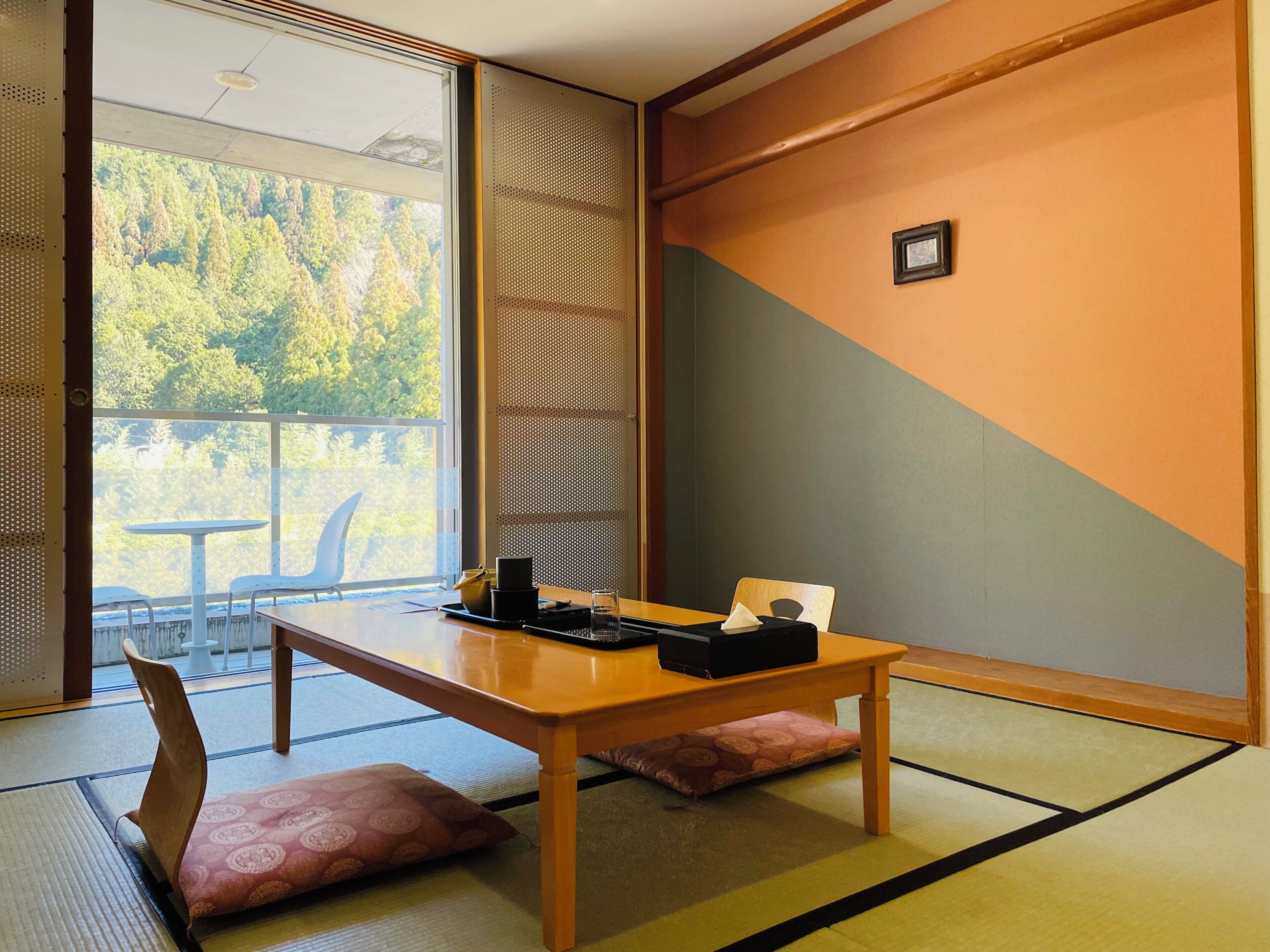 A traditional Japanese room with tatami flooring, a low table with a black tray, a wooden chair, and a sliding door leading to a balcony with a view of trees.