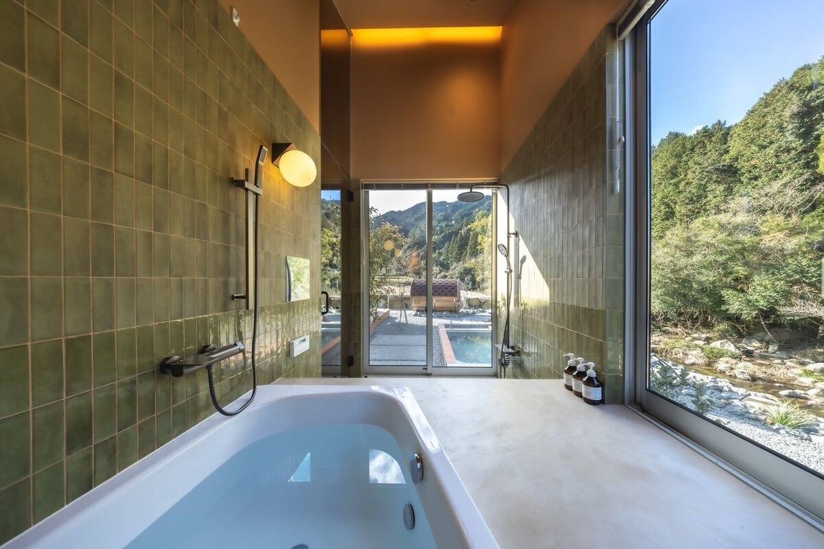 A modern bathroom with a large bathtub, a shower, and a view of a natural landscape through a glass door.