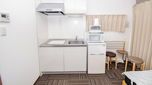 Una kitchenette compacta con microondas, lavabo y armarios, una pequeña mesa con dos taburetes y una cama con colcha a rayas.