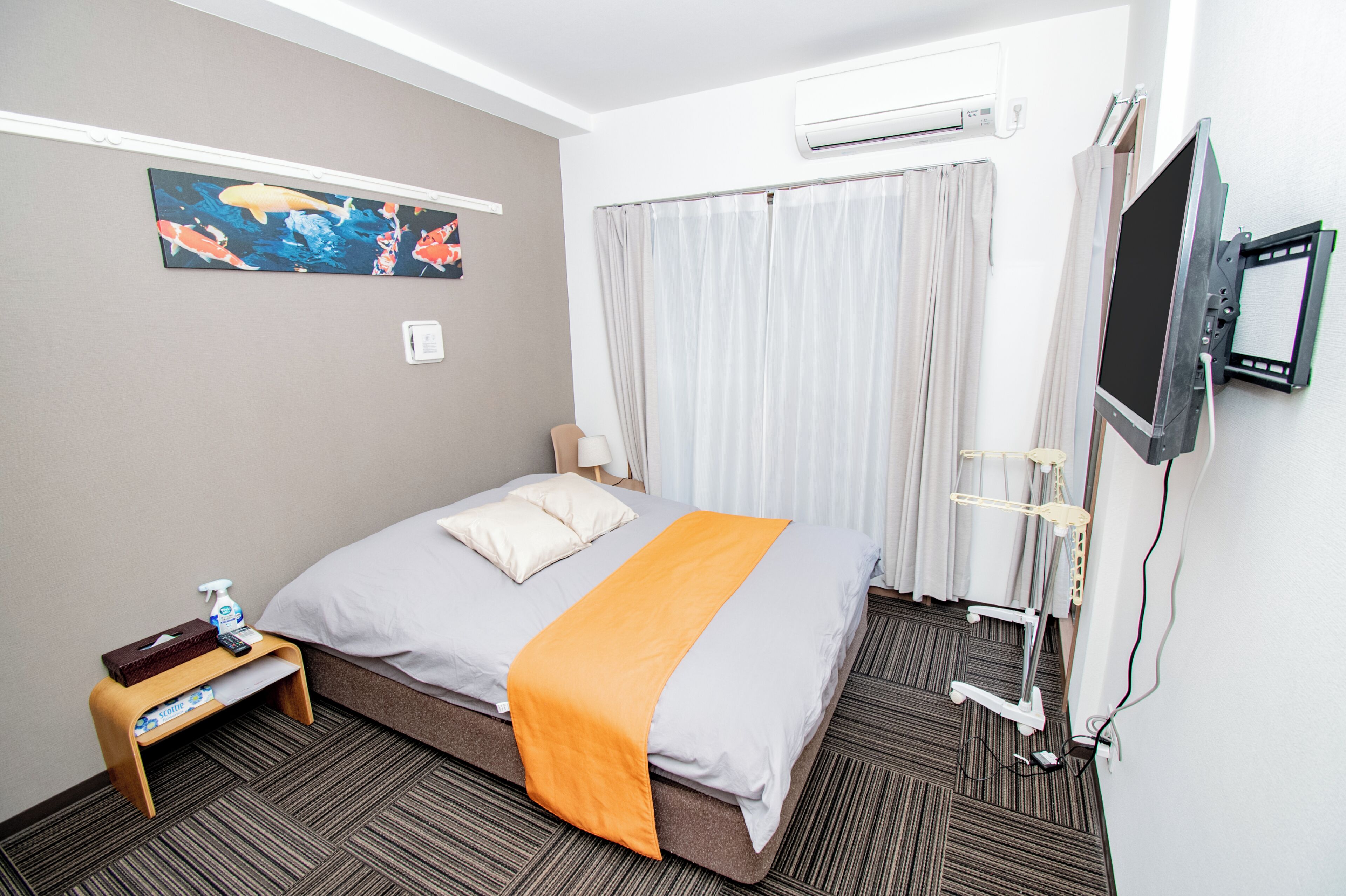 A hotel room with a bed, an air conditioning unit, a television, a small table with a tissue box and spray bottle, and a painting on the wall.