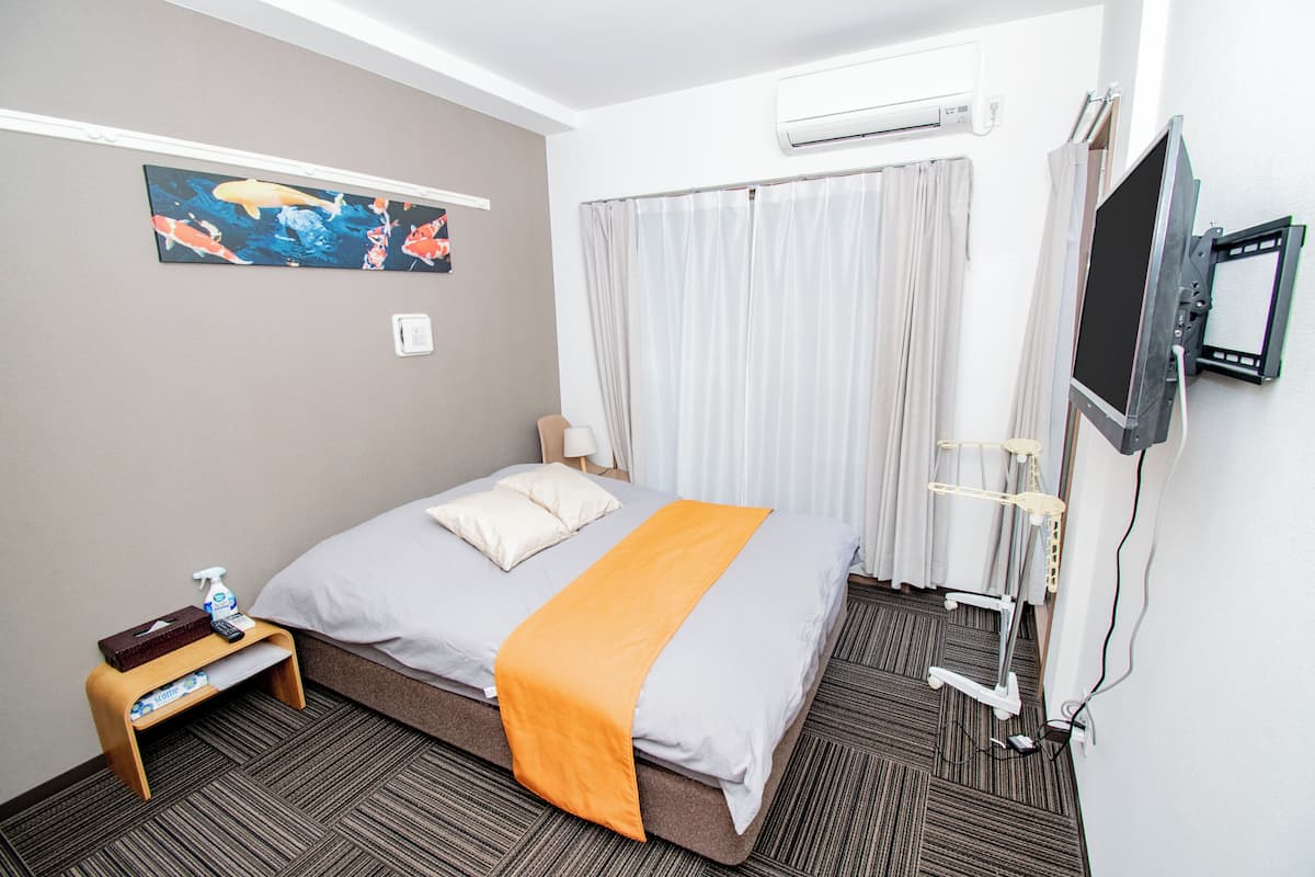 Ett hotellrum med en säng, en luftkonditioneringsanläggning, en tv, ett litet bord med en näsdukask och en sprayflaska samt en tavla på väggen.