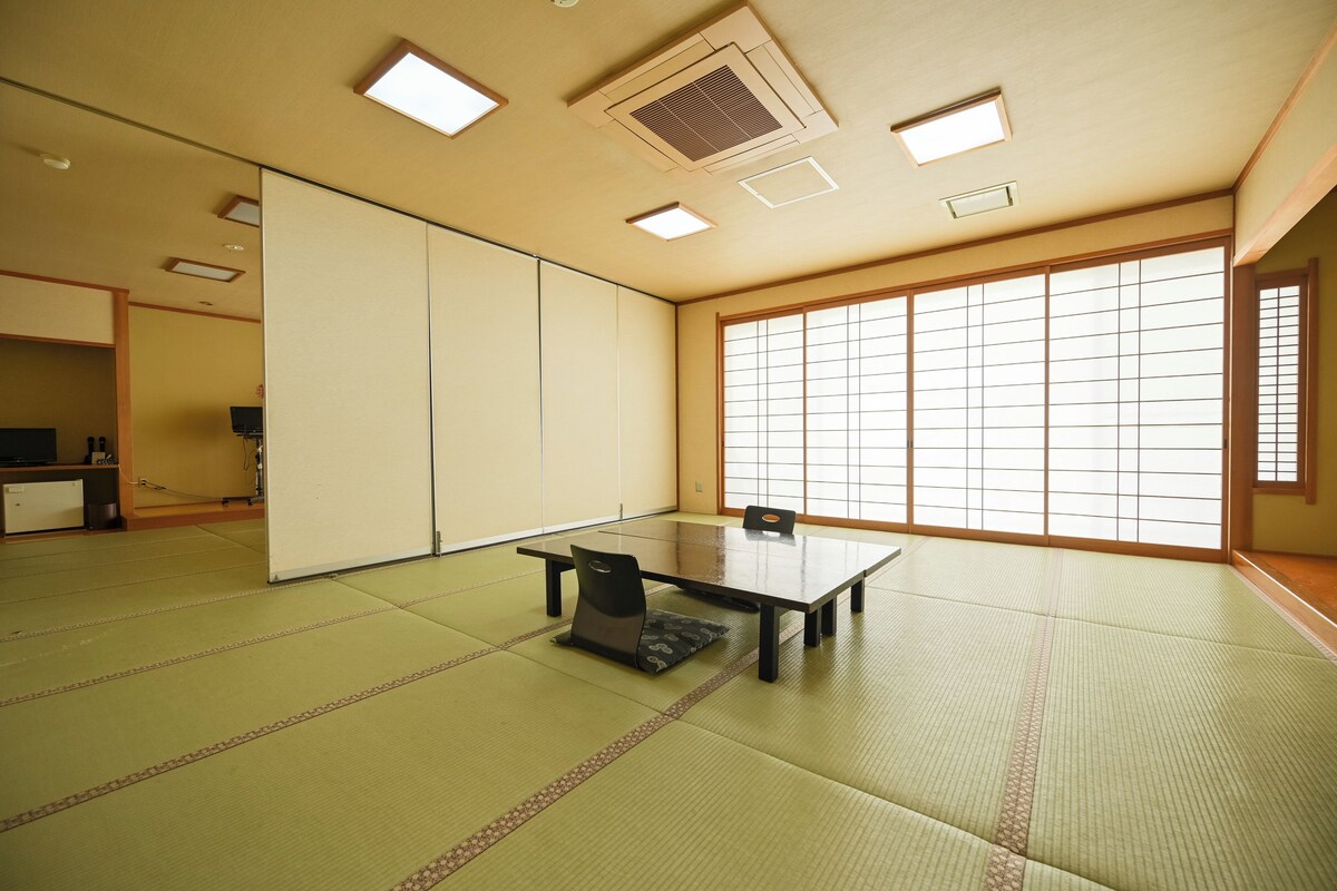 Une pièce avec un sol recouvert de tatamis, une petite table, des chaises, des portes coulissantes et de grandes fenêtres.