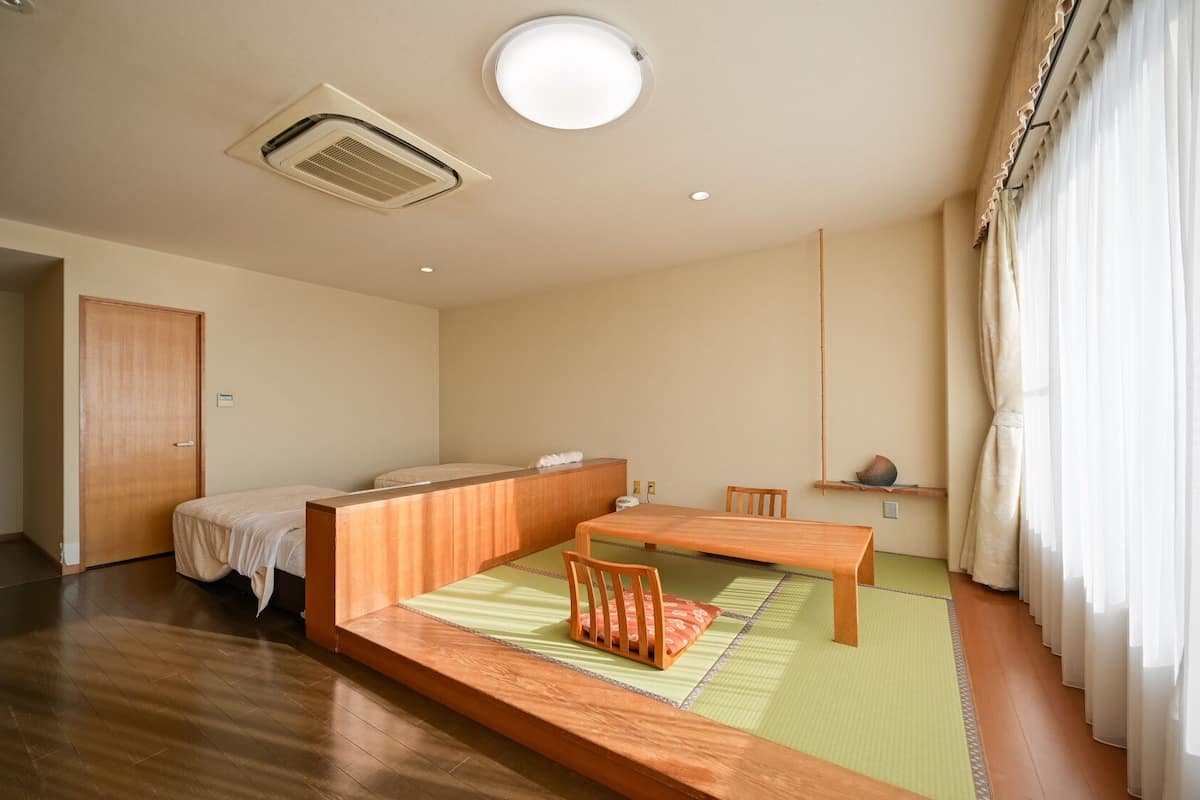 Una habitación con cama y mesa de madera, esteras de tatami, un ventilador de techo y una ventana con cortinas.