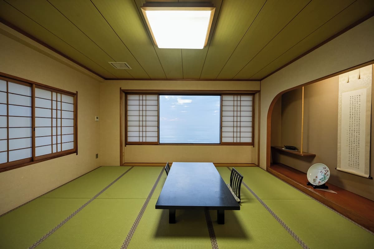 Una habitación con esteras de tatami, una mesa de tatami, puertas correderas, un estante con un cuenco y un pergamino en la pared.