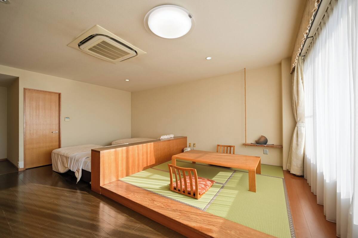 Una habitación con una cama, una mesa de madera, una silla, un futón, una ventana con cortinas y un respiradero en el techo.