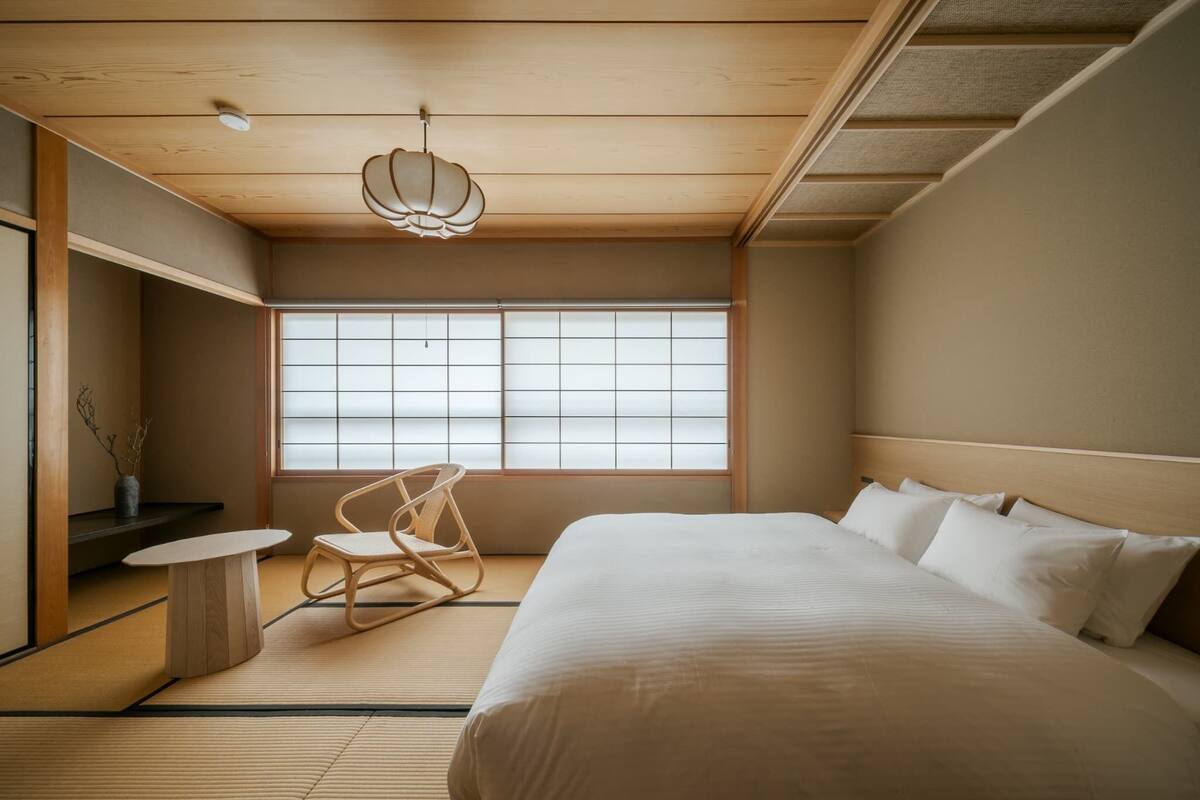 A minimalist Japanese-style bedroom with tatami flooring, a low wooden bed, a traditional chair, and a large window with shoji screens.