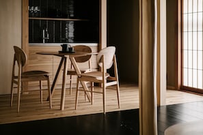 A minimalist dining area with a round table and four chairs, a window with blinds, and a kitchen area in the background. - Anjin Gosho Ebisugawa (Kyoto)