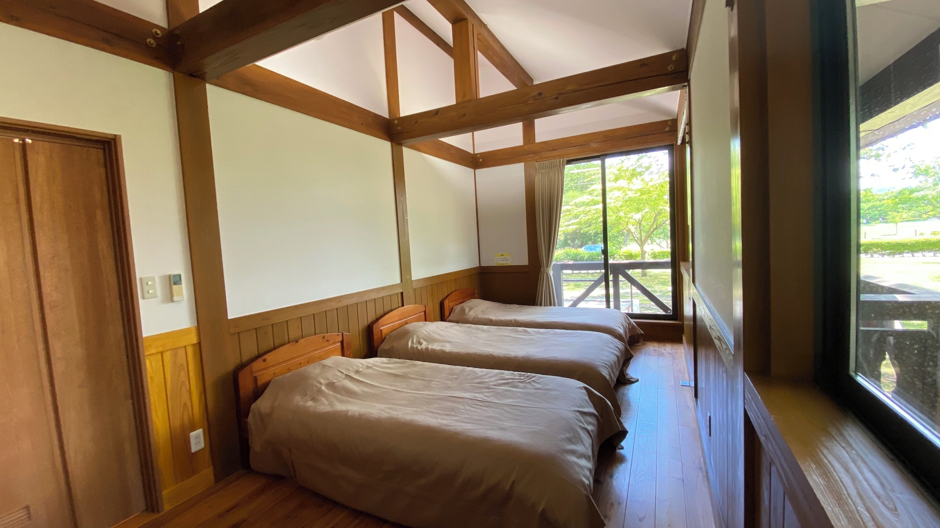 A room with wooden beams, two beds, a window with a view of greenery, and a door.