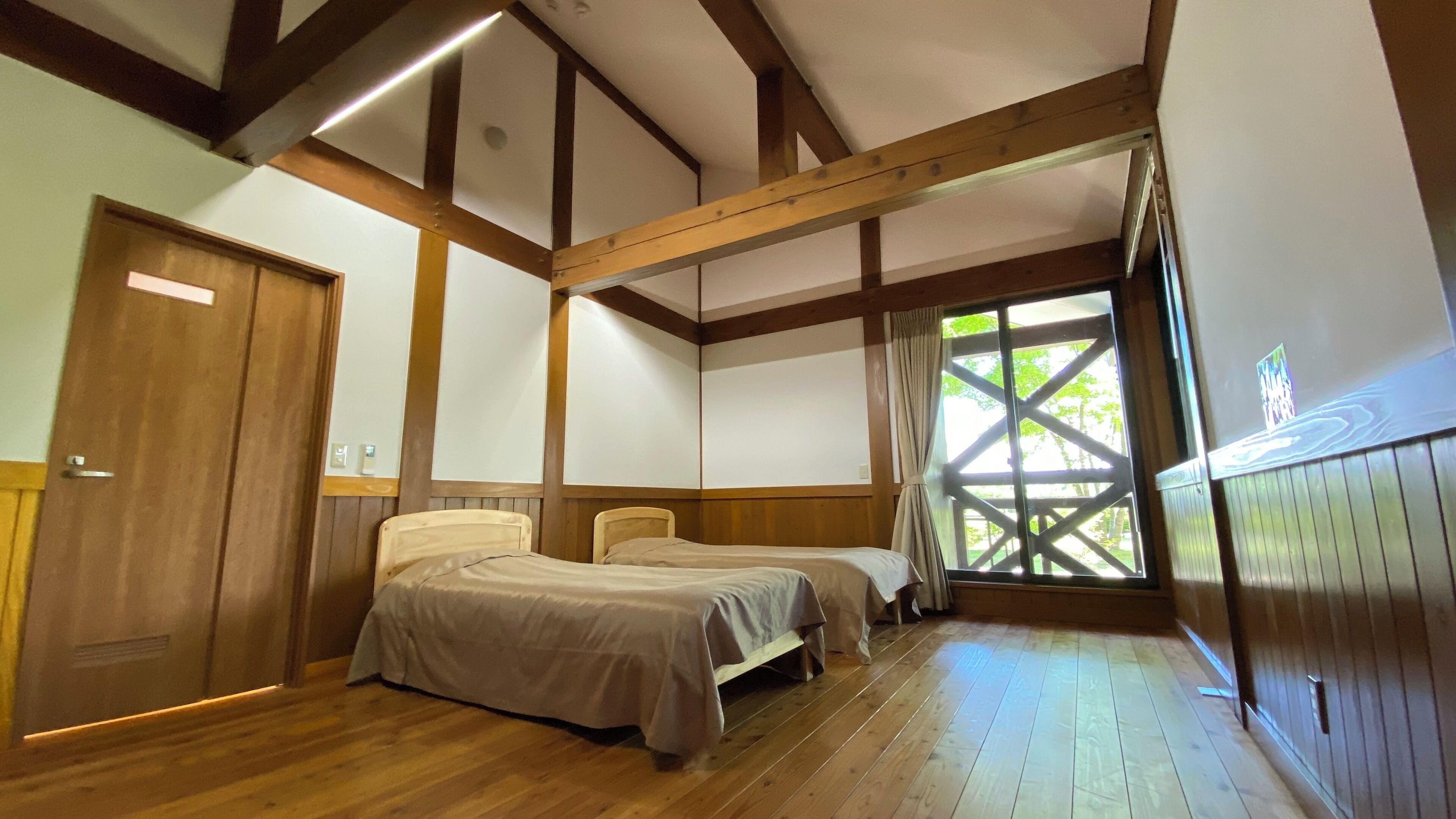 A room with two beds, wooden beams, a large window with a view, and a door.
