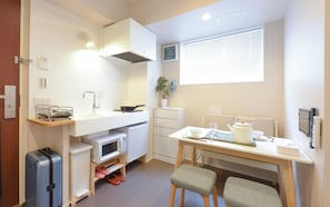Een kleine keuken met een magnetron, fornuis en spoelbak, een eettafel met stoelen, en een koffer.