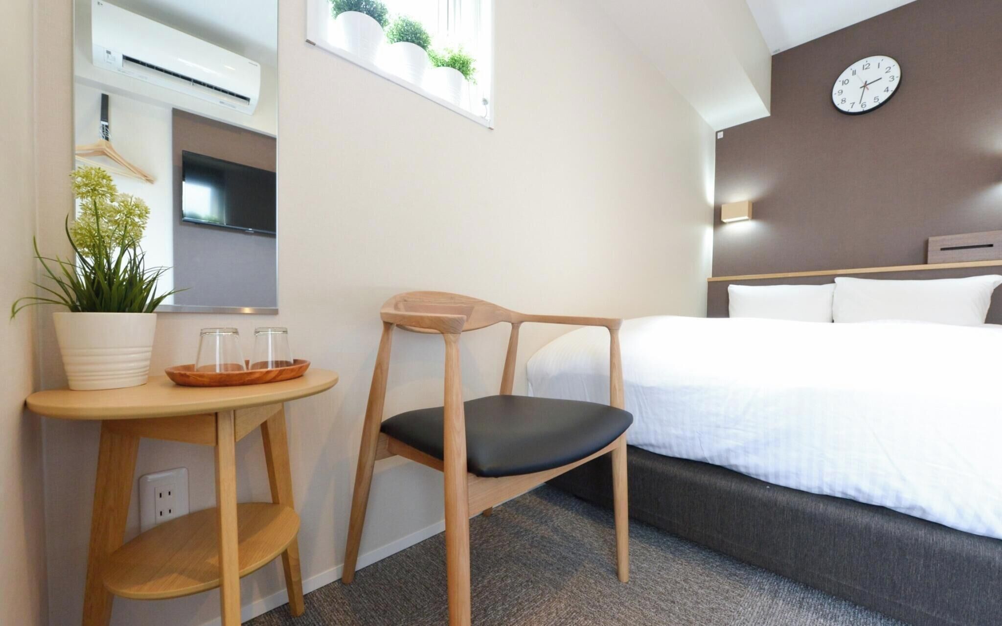 A hotel room with a bed, a wooden chair, a small table with a plant, a TV, and a wall clock.