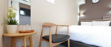 A hotel room with a bed, a desk with a chair, a potted plant, a clock on the wall, and an air conditioning unit.