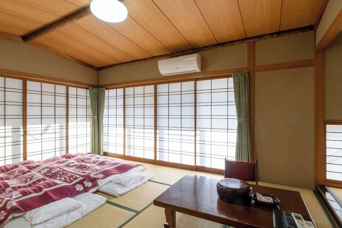A traditional Japanese room with tatami flooring, a futon bed, a shoji screen, a wooden table with a kettle, and an air conditioning unit.
